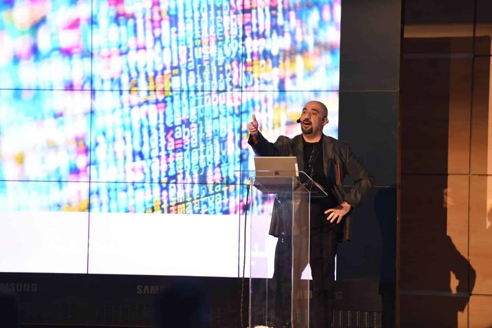 Dr. Mohamed Qasem speaking on stage before a wall of code